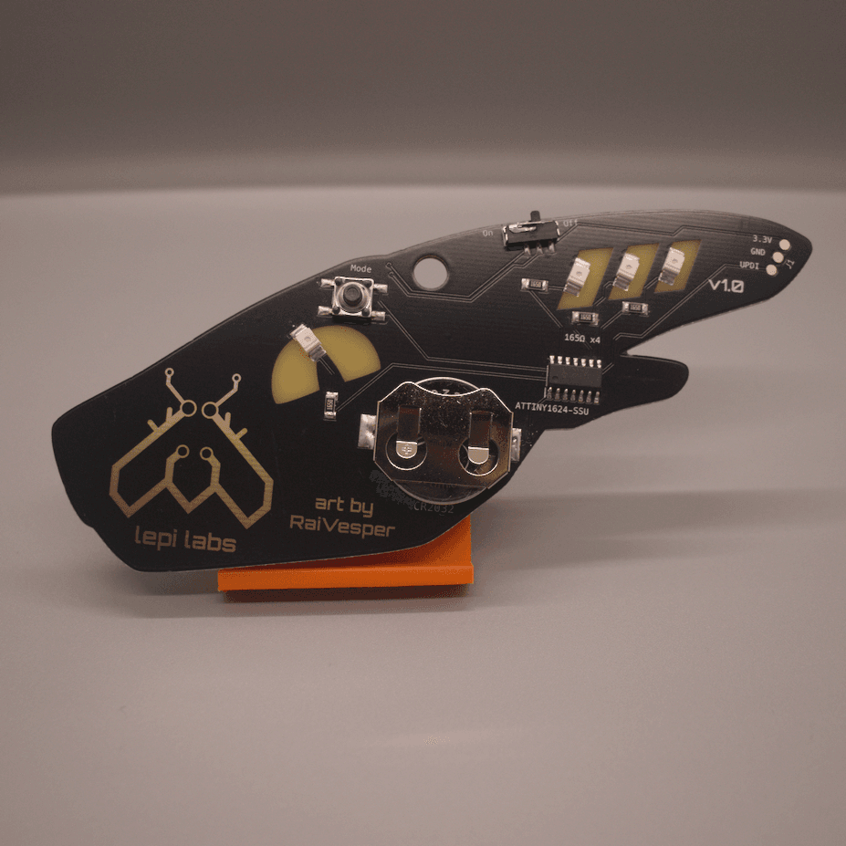 Photo of the rear view of the synth PCB badge. The components shown include a push button to change modes, three reverse mounted LEDs, a CR2032 coin cell battery holder, a microcontroller, a sliding power switch, and a connector at the tip for UPDI programming.