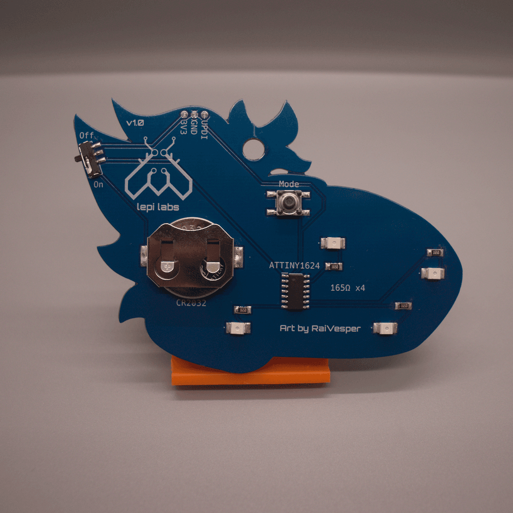 Rear view of a blue protogen badge, showing the components which include LEDs, resistors, a coin cell battery, a microcontroller, a push button mode switch and a sliding power switch, and a UPDI connector.